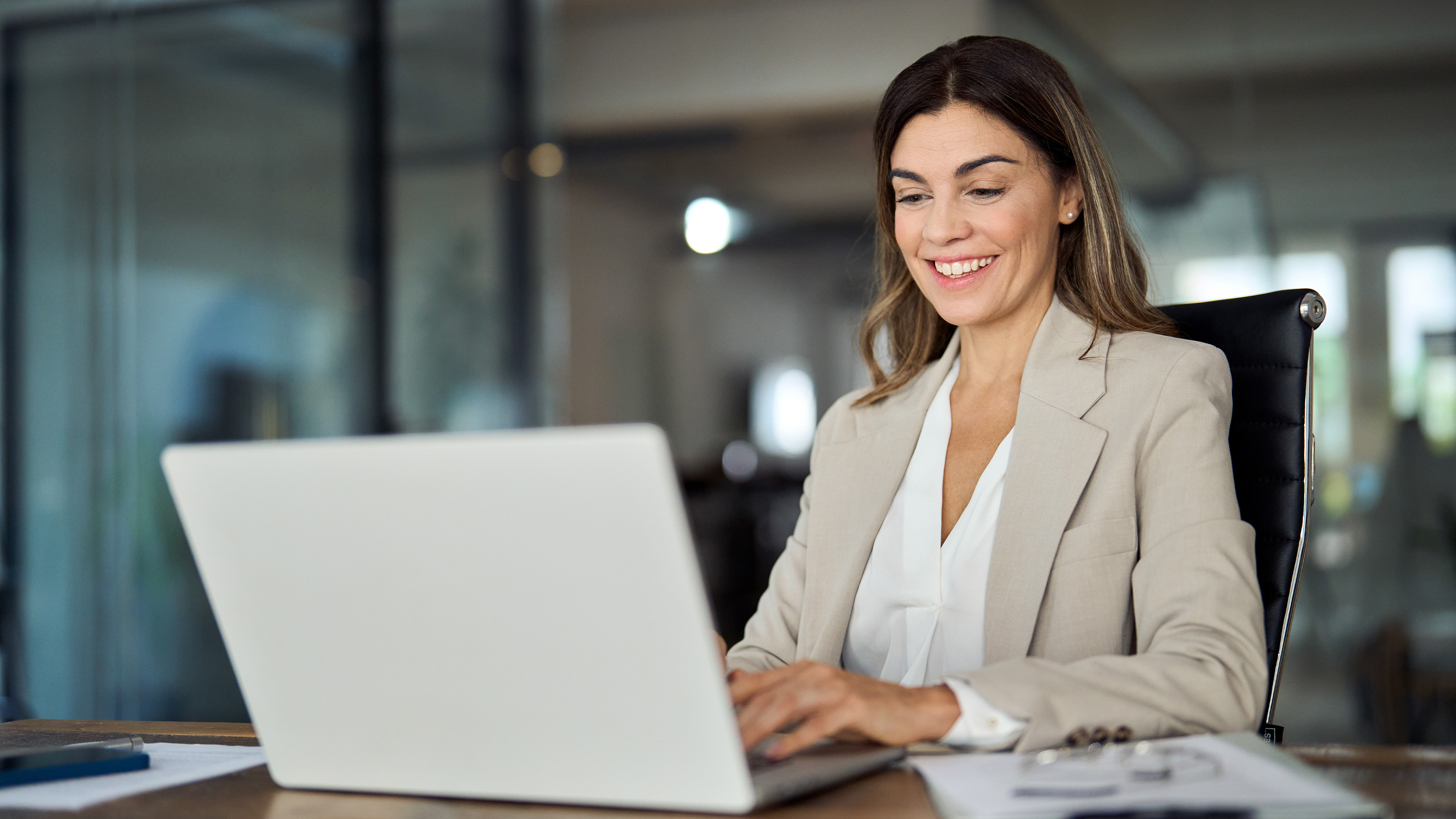 Smiling businesswoman in a beige blazer typing on a laptop at an office desk