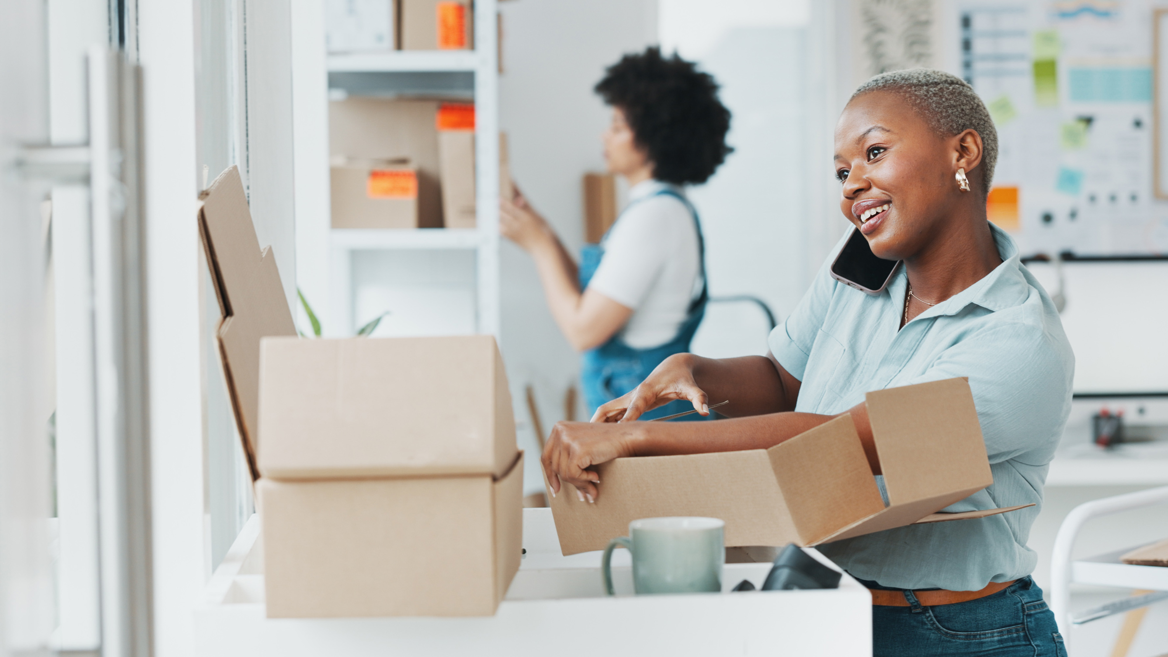 Smiling woman on a phone call unpacking a moving box in an office while a coworker organizes shelves in the background