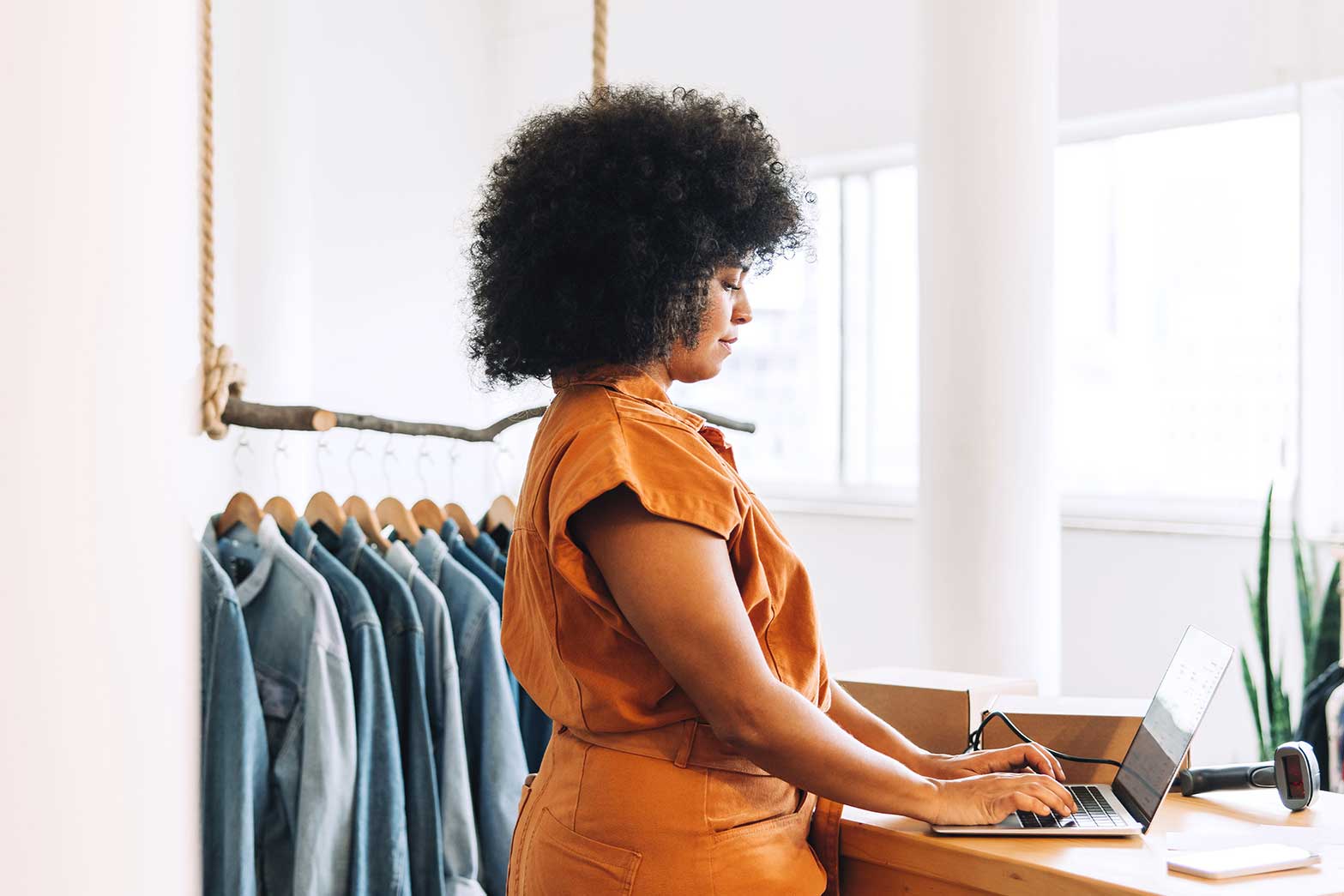 Woman in an orange outfit typing on a laptop at a counter beside a rack of hanging denim in a bright boutique.