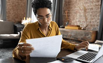 Person in glasses reviewing paperwork at a desk with a laptop and calculator