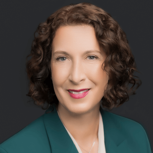 Headshot of Pachuta, a woman with curly brown hair, smiling in a teal blazer against a dark background.