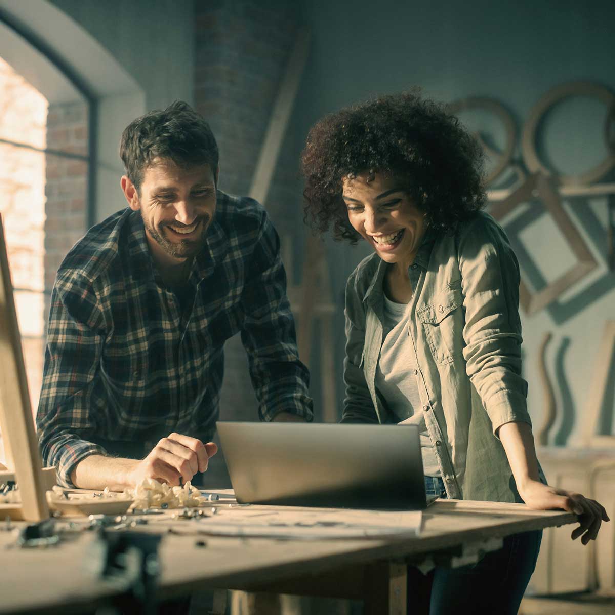 Man and woman smiling while looking at a laptop together at a workbench in a woodworking studio.
