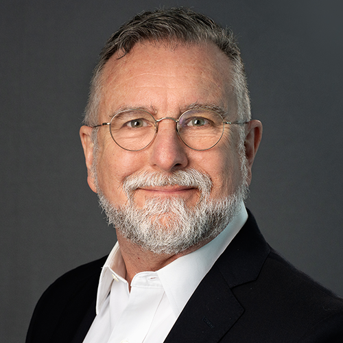 Headshot of Gillespie, a man with a gray beard and round glasses, smiling in a dark suit and white shirt.