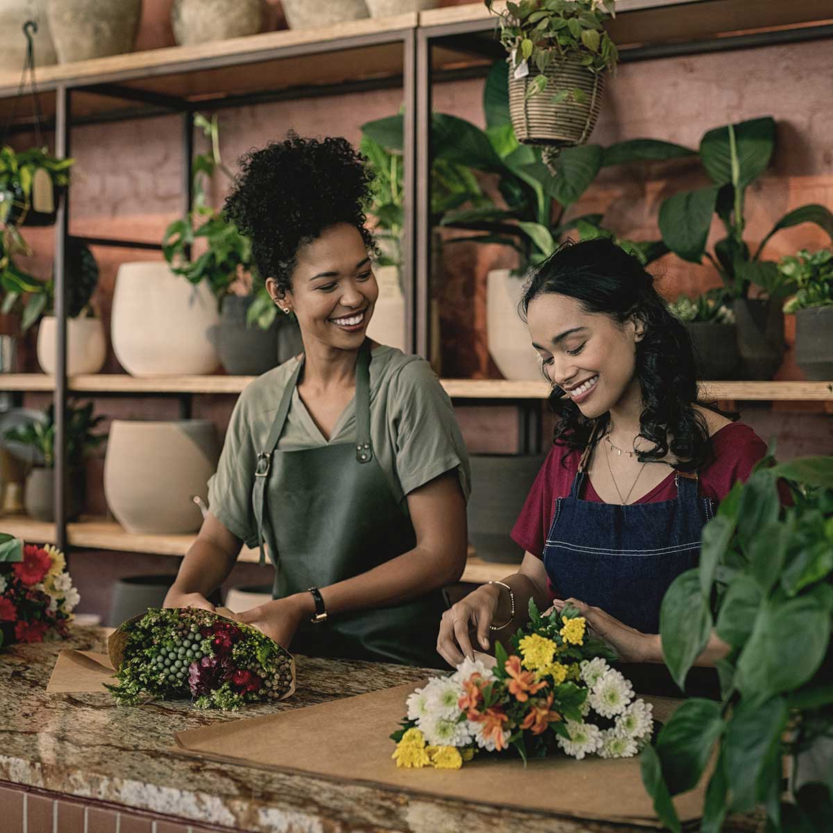 Two smiling women arranging flower bouquets at a counter in a plant shop lined with pots and greenery.