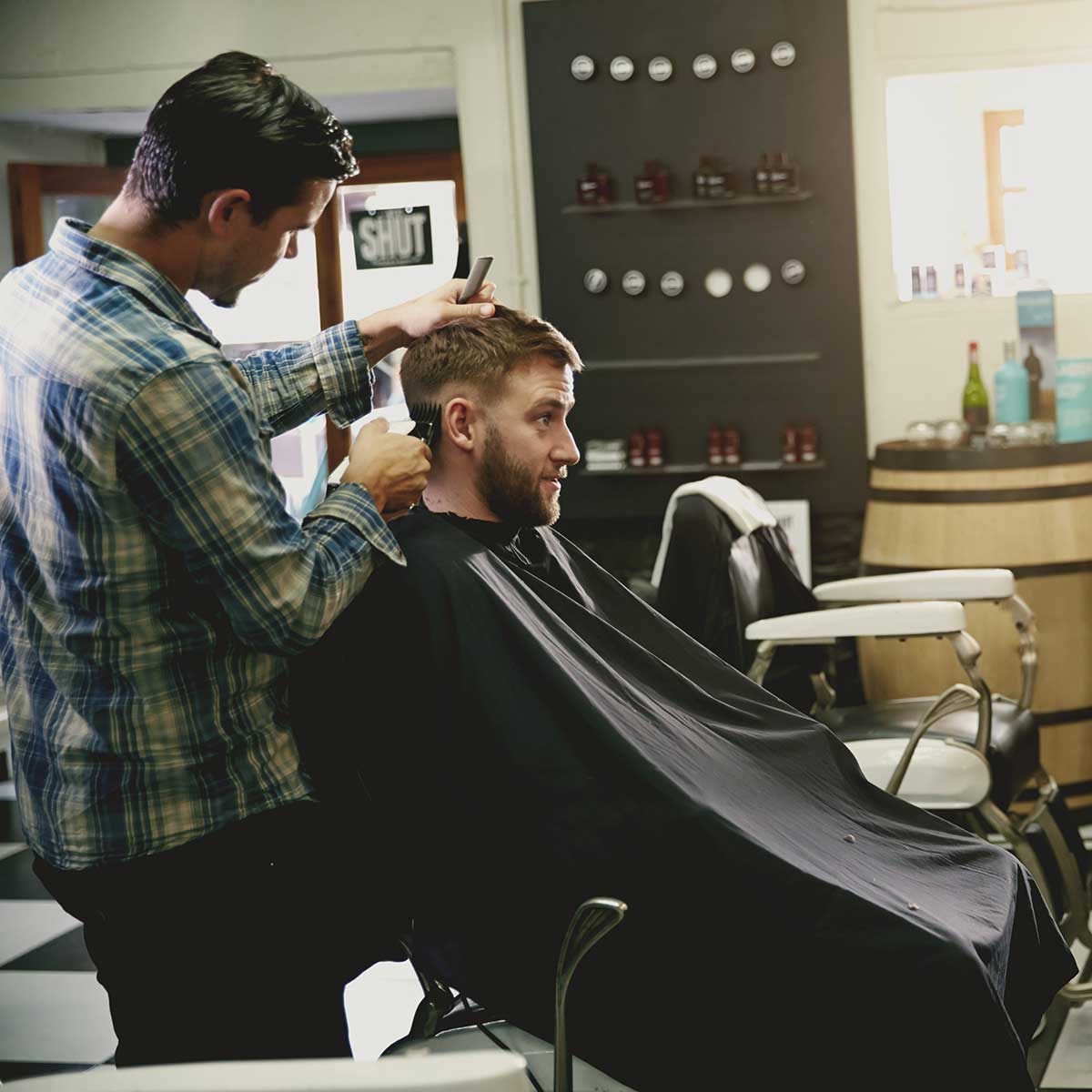 Barber in a plaid shirt cutting a seated client's hair in a barbershop with product shelves in the background.