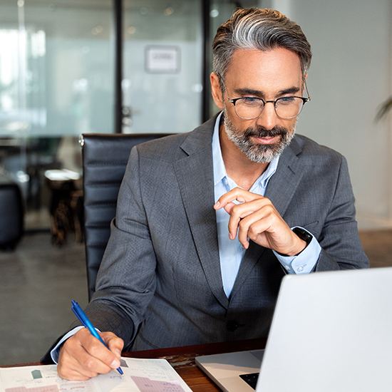 A businessman in a suit smiling while writing notes at his desk next to a laptop