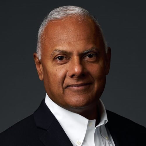 Headshot of Raghav Nandagopal, a man with gray hair, smiling in a dark suit and white shirt against a dark background.