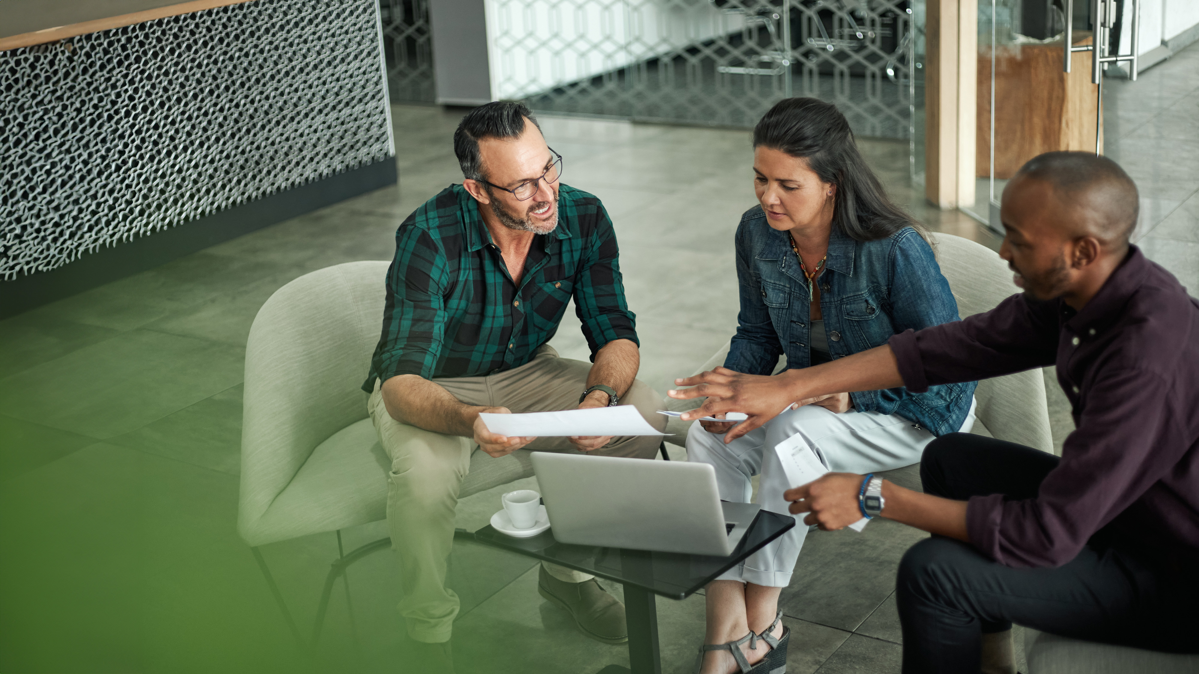 Three colleagues discussing paperwork together on lounge chairs in an office lobby, with a laptop on the table