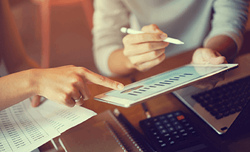 Hands reviewing financial charts on a tablet alongside printed reports and a calculator