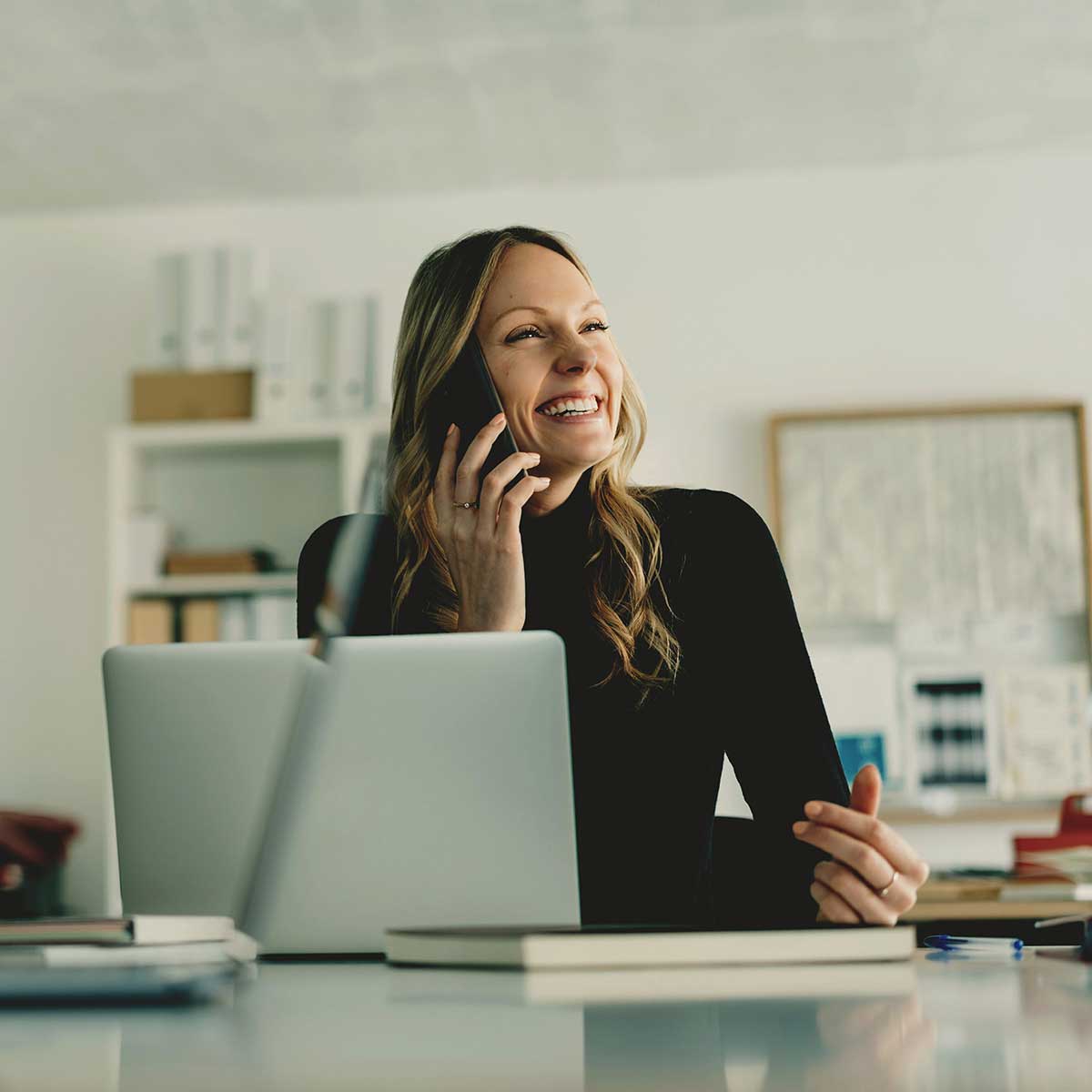 Woman in a black top laughing while talking on her phone at a desk with a laptop and notebooks.