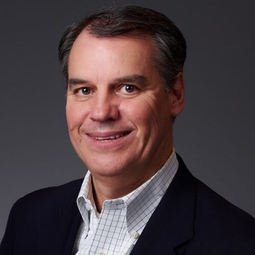 Headshot of Jim Gearin smiling in a dark suit with a checkered shirt against a dark background.