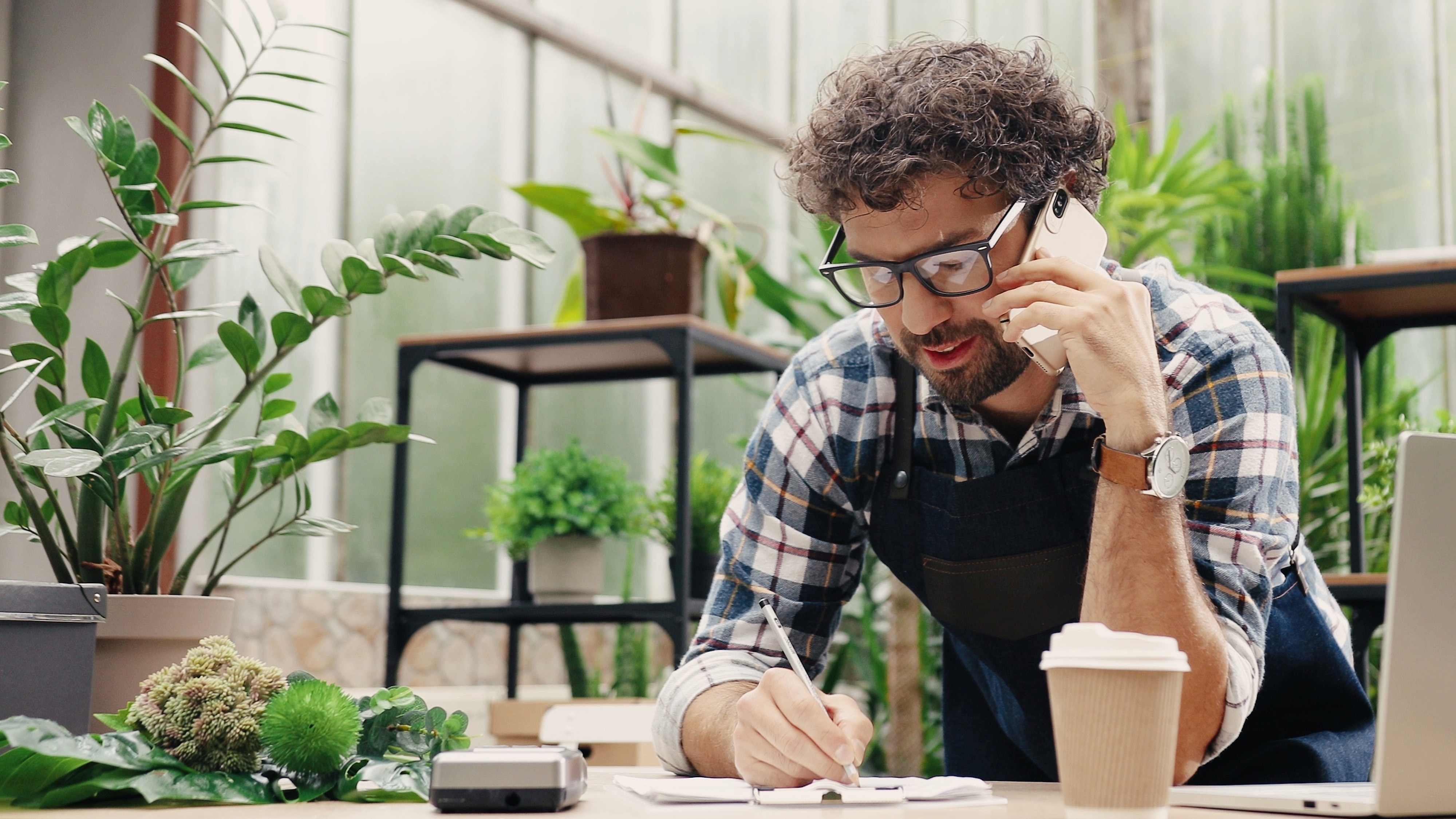 Plant shop owner on the phone writing notes on a clipboard surrounded by greenery