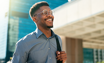 A smiling young man wearing glasses and carrying a bag walking outdoors near an office building.