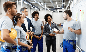 A group of warehouse workers wearing hard hats listening to a colleague present at a whiteboard