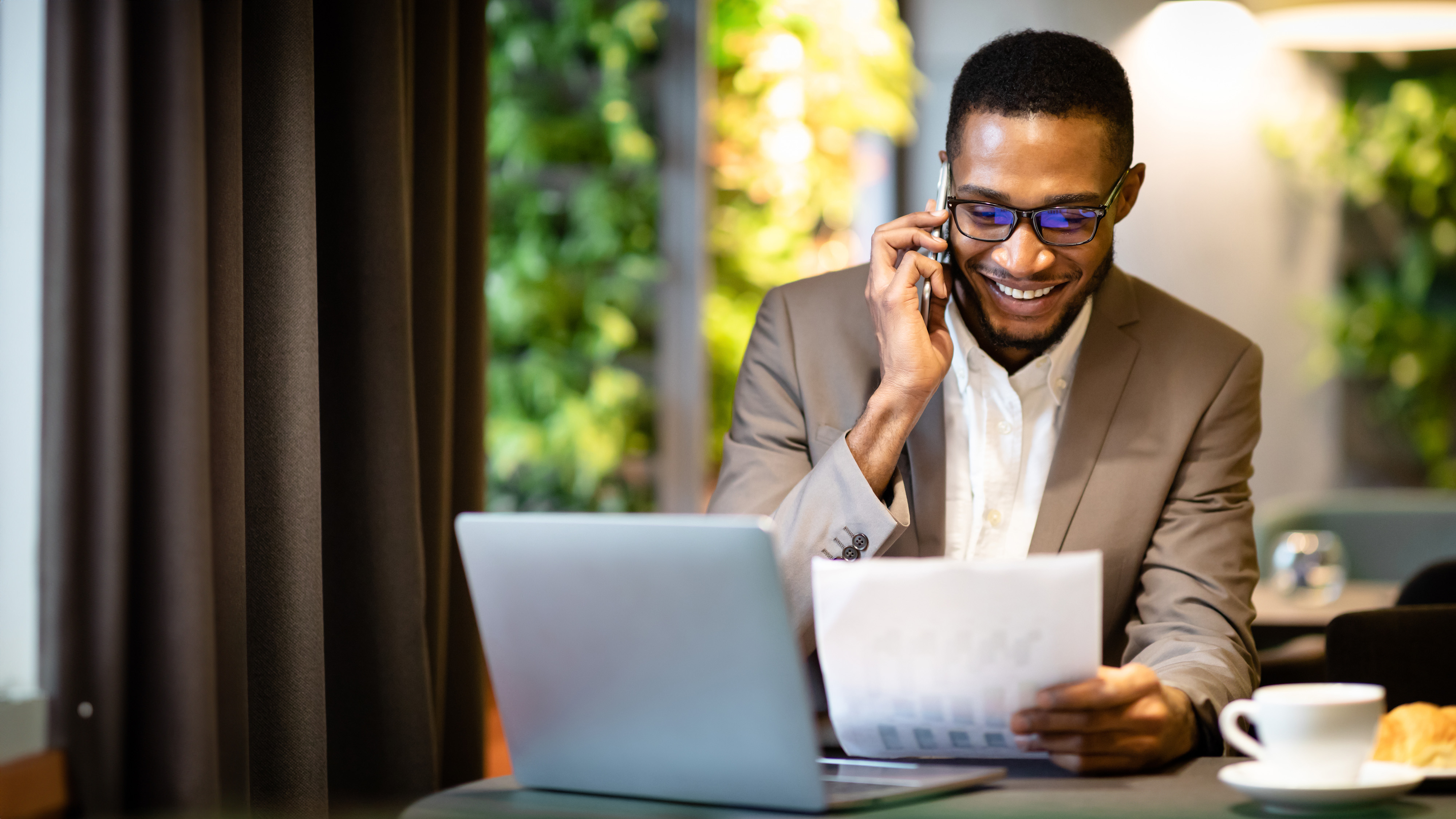 Businessman on a phone call reviewing a financial chart printout at a cafe table with a laptop