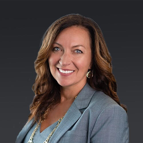 Headshot of Kasey Price smiling with long wavy brown hair, wearing a gray blazer against a dark background.