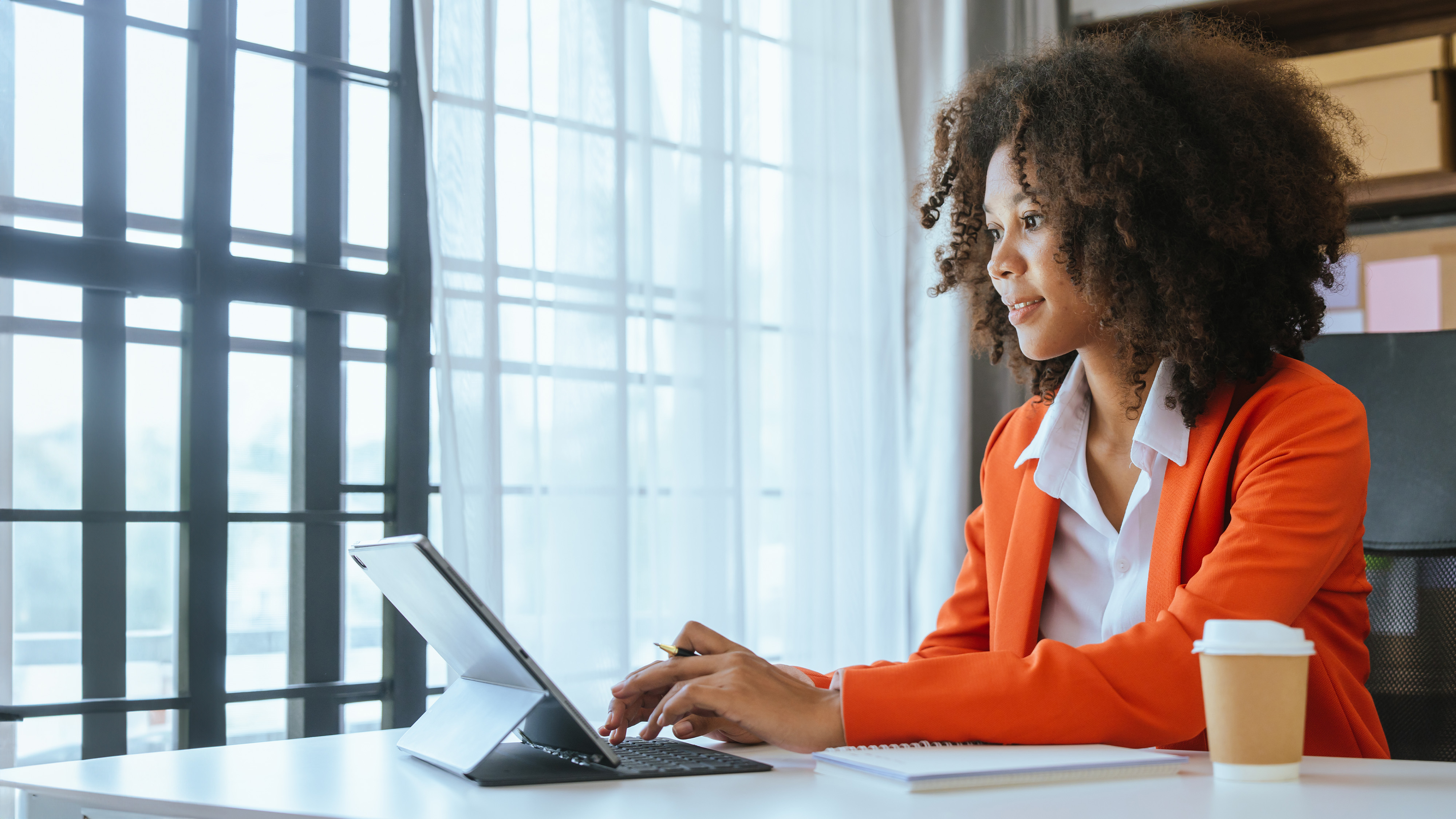 Businesswoman in an orange blazer working on a tablet at a desk near a window