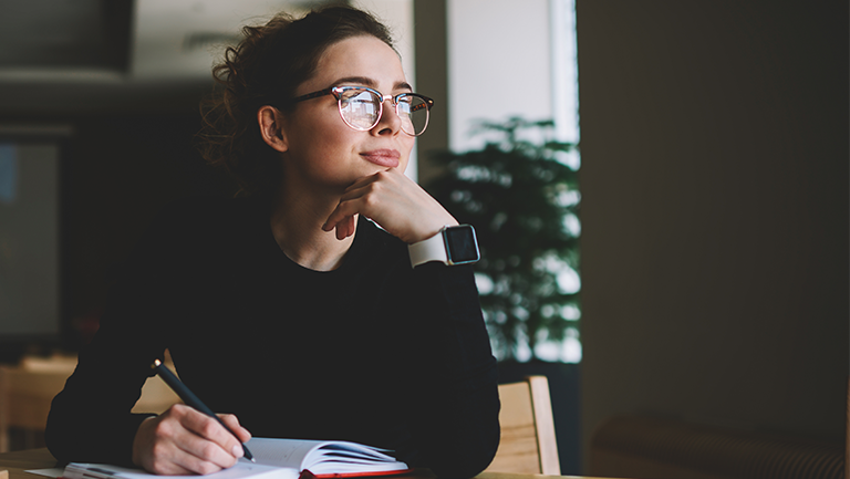 Woman with glasses pausing thoughtfully while writing in a notebook at a desk
