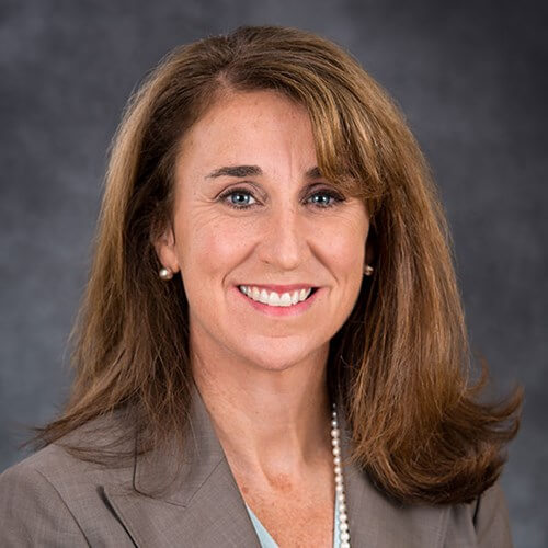 Headshot of Peg Creonte smiling in a taupe blazer with a pearl necklace against a dark gray background.